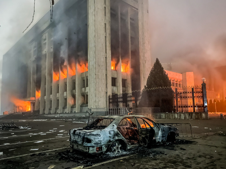 Egy tüntetők által felgyújtott és kiégett autó, a háttérben a lángokban álló polgármesteri hivatallal, alatta pedig a hivatal épületében elkövetett rongálás nyomai a legnépesebb kazah városban, Almatiban 2022. január 5-én – Fotó: Valery Sharifulin / TASS / Getty Images; Stringer / Reuters