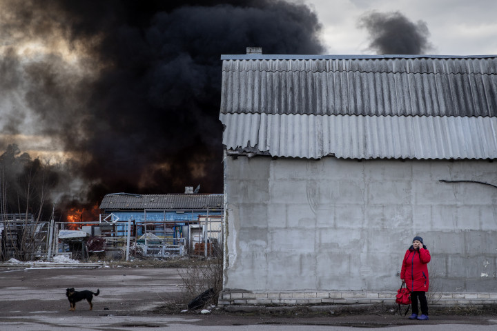 Egy nő keres menedéket egy épület mögött, miközben füst száll fel egy vegyianyag-raktárból, amit orosz támadást ért az ukrajnai Kalinivkában 2022. március 8-án – Fotó: Chris McGrath / Getty Images