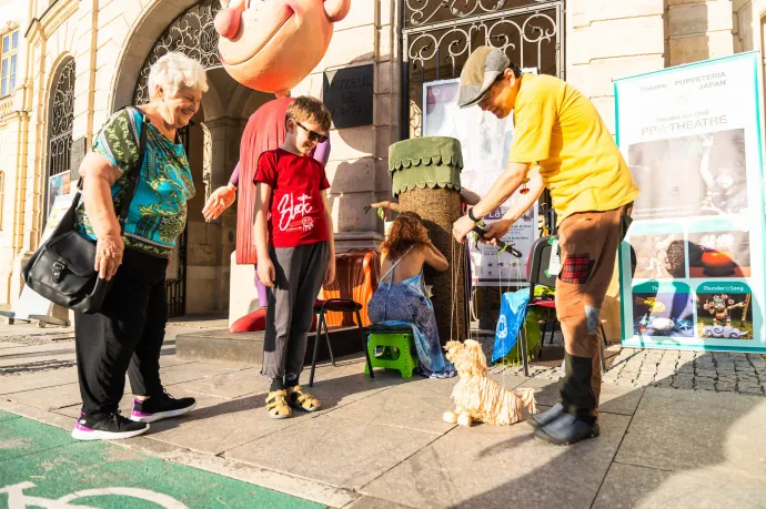 Kaz a pulikutya báb mozgatásával szórakoztatja a nézőket, miközben a háttérben a fatörzs elé telepedett néző megtekinti a Yuki által előadott háromperces produkciót – Fotó: Wonder Puck Fesztivál hivatalos Facebook-oldala