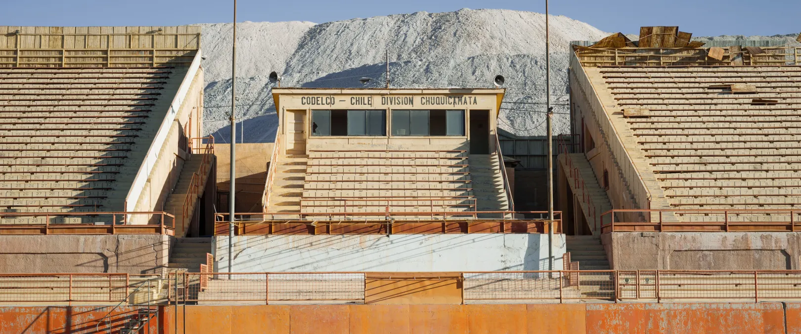 Chuquicamata szellemvárosa a hozzá tartozó üresen kongó stadionnal. A 2004 és 2007 között kiürített várost egy egykori bányásztábor helyén építették fel, de ahogy egyre nagyobb területet foglaltak el a külszíni fejtések úgy lehetetlenült el a település. A feldolgozóegységekből ontott mérgező gázok és por miatt végül elköltöztették az egész várost. Egy kllogramm rézérc kibányászása száz kllogrammnyi meddőanyag fejtésével jár, a meddő egy részét pedig a településen terítették szét – Fotó: Davide Monteleone