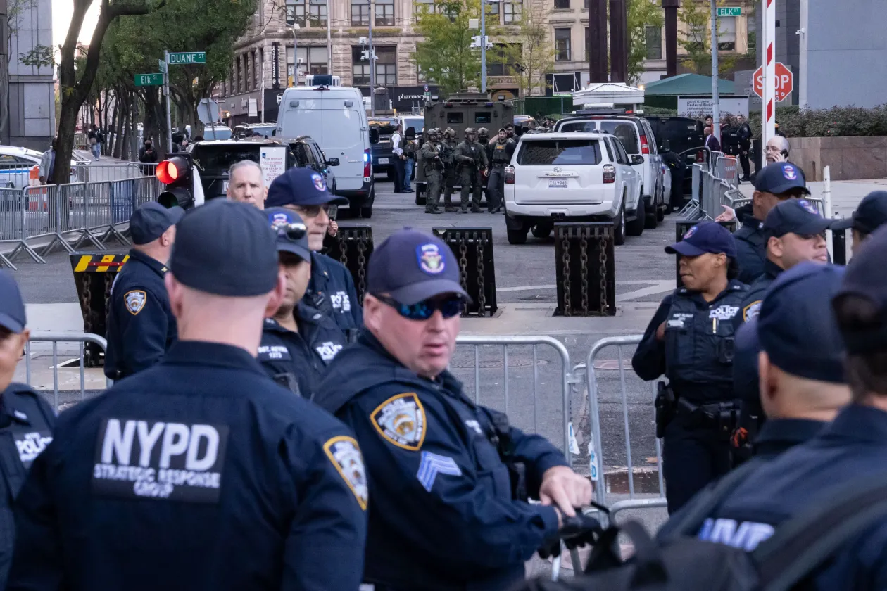 Állig felfegyverzett szövetségi ügynökök strázsálnak egy BearCat páncélozott szállítójármű mellett, az előtérben New York-i rendőrök; a jobboldali képen pedig a belbiztonsági minisztérium egyik bevetési parancsnoka, a posztjához képest igen laza szerelést viselő William Joyce int a fotósnak egy 2025. október 21-én végrehajtott New York-i akció során – Fotó: Adam Gray / Getty Images