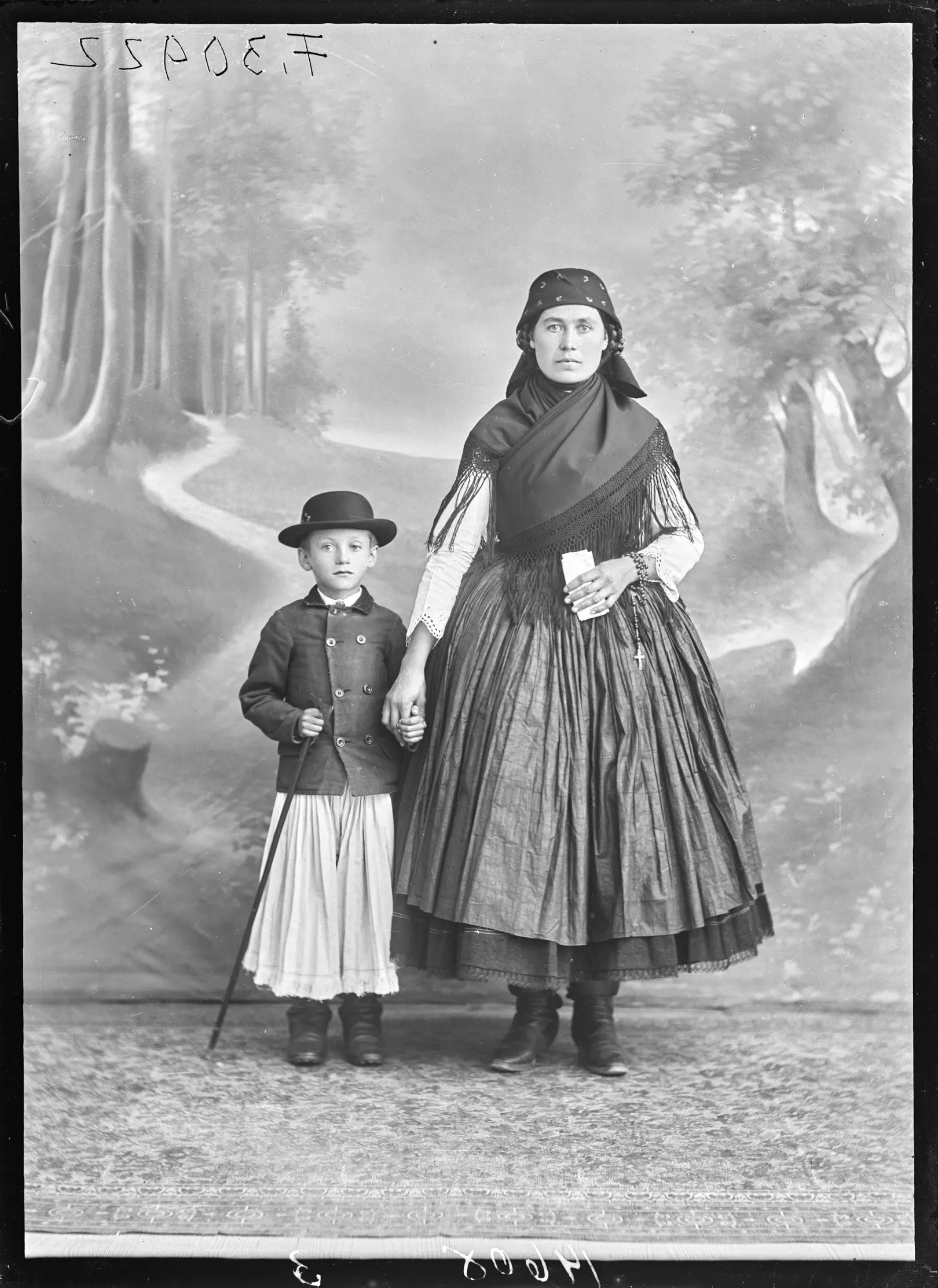 Palóc asszony kisfiával, erdőrészletet és benne futó utat ábrázoló festett háttér előtt, Eger, 1910-es évek – Fotó: Néprajzi Múzeum Fényképtára / Kiss József felvétele