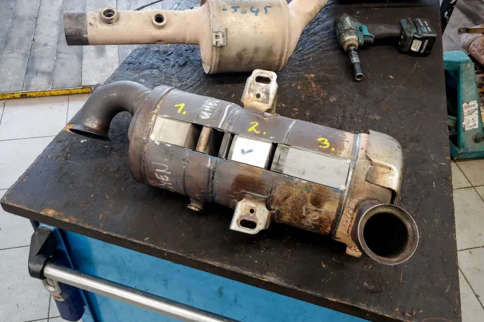 Fent Varga József és Varga Viktor, a háttérben az általuk konstruált részecskeszűrő- és redukcióskatalizátor-mosó berendezéssel, kezükben egy Stellantis-féle 1,5 literes BlueHDI dízelmotor szétvágott oxidációs, illetve redukciós katalizátort, valamint részecskeszűrőt is tartalmazó egysége, ami a lenti képen is látható – Fotó: Zách Dániel / Telex