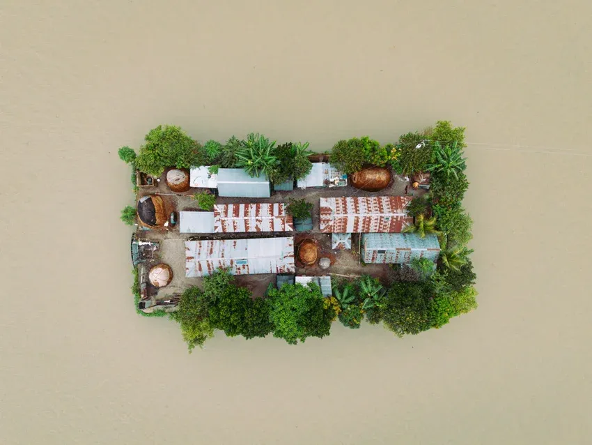 Az egymástól elvágott, szigetszerű településeken házakat és haszonállatokat is látni. A természetes magaslat kilátszik a monszun miatti árvíz alól. / Ashtagram egyetlen megemelt útja köti össze a magasházakat. Az árvízben látszanak az elektromos hálózat póznái – Fotó: Joy Saha