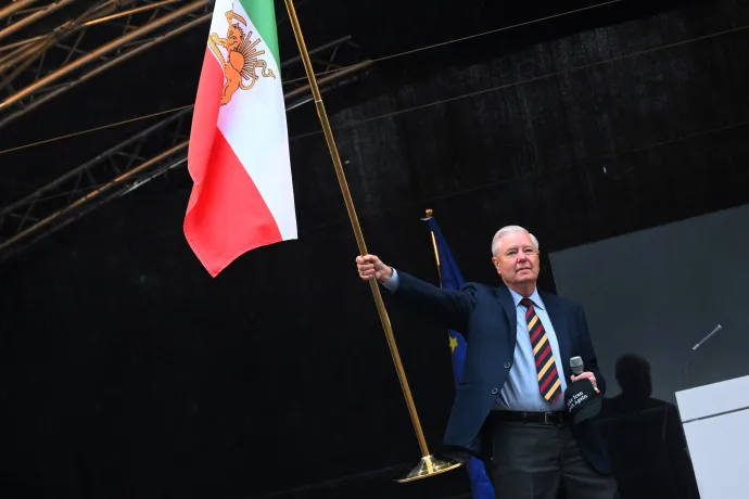 Lindsey Graham amerikai politikus az 1979 előtti iráni zászlót tartva áll egy, az „Emberi jogok és szabadság Iránban: nemzetközi szolidaritás az iráni néppel” címmel tartott tüntetésen Münchenben, a Müncheni Biztonsági Konferencia ideje alatt 2026 februárjában – Fotó: Felix Hörhager / picture alliance / Getty Images